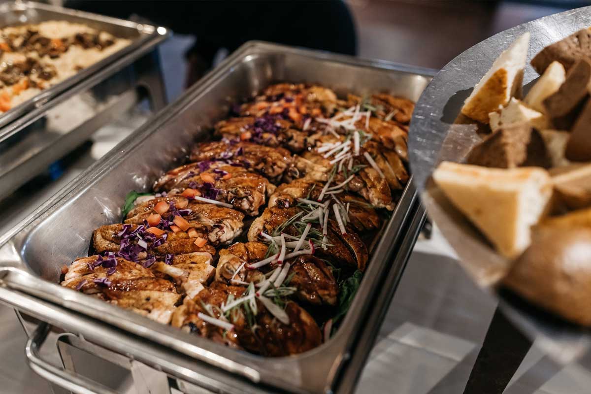 A metal buffet tray filled with cooked chicken breast topped with vegetables and herbs, with a side dish visible on the right.