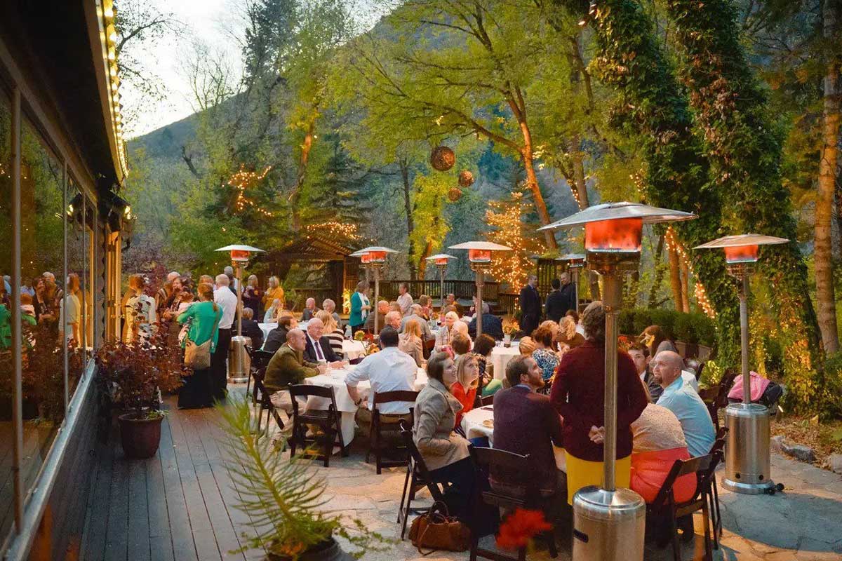 A group of people seated at outdoor tables, dining and socializing at an evening event surrounded by greenery and string lights.