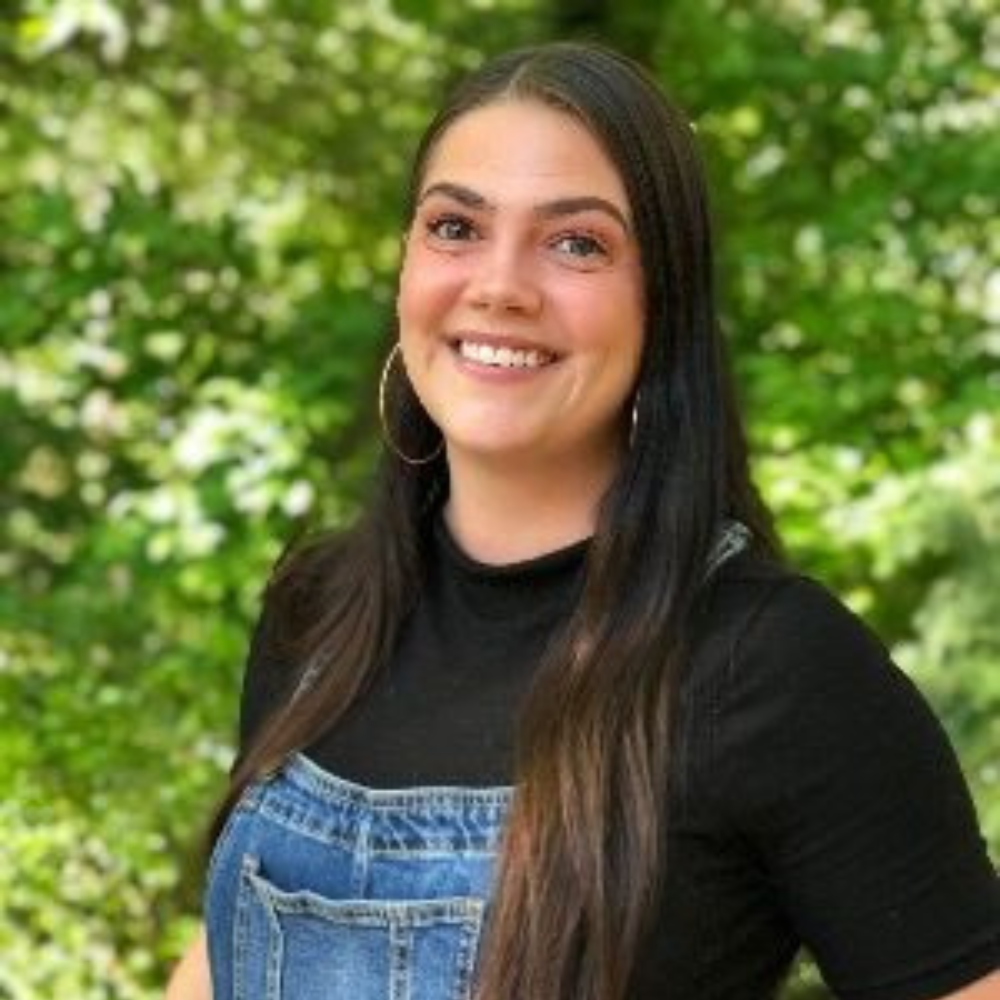 A woman with long dark hair wearing a black top and denim overalls smiles outdoors with a green leafy background.