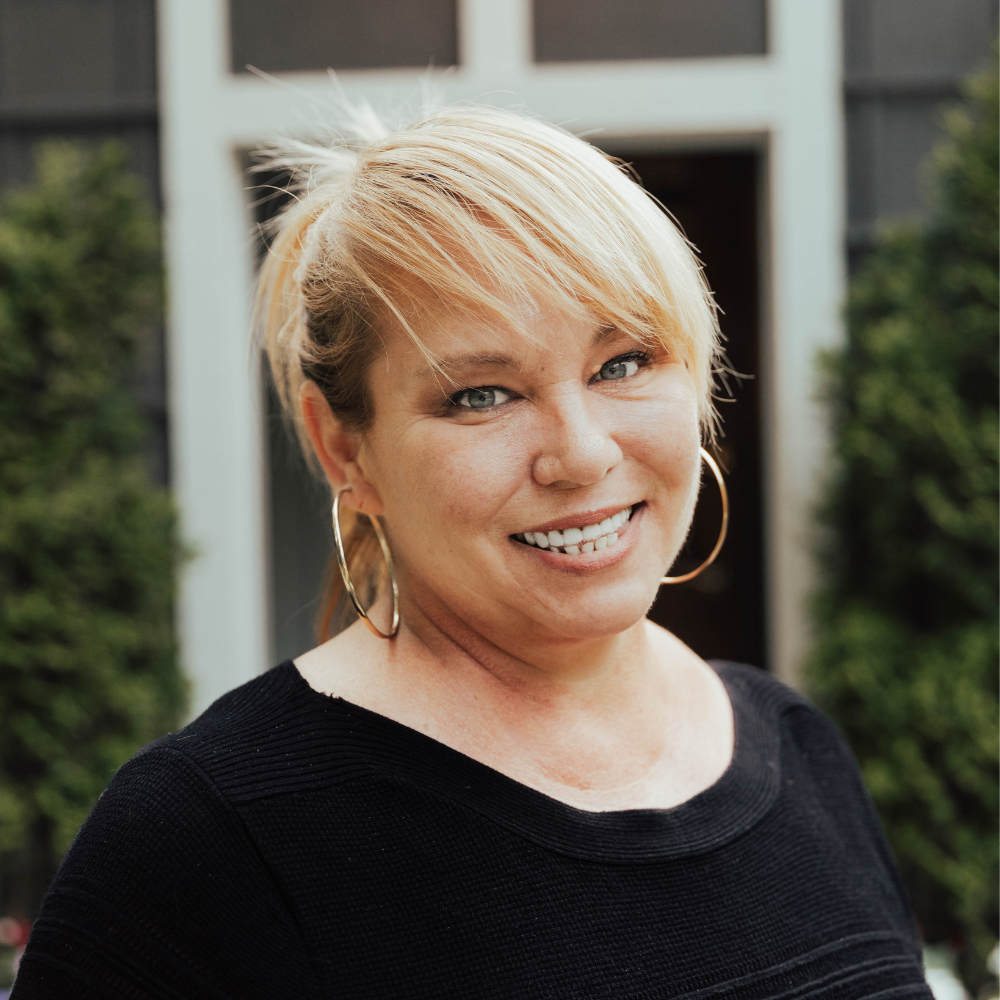 A woman with blonde hair, wearing large hoop earrings and a black top, smiles in front of a door and some greenery.